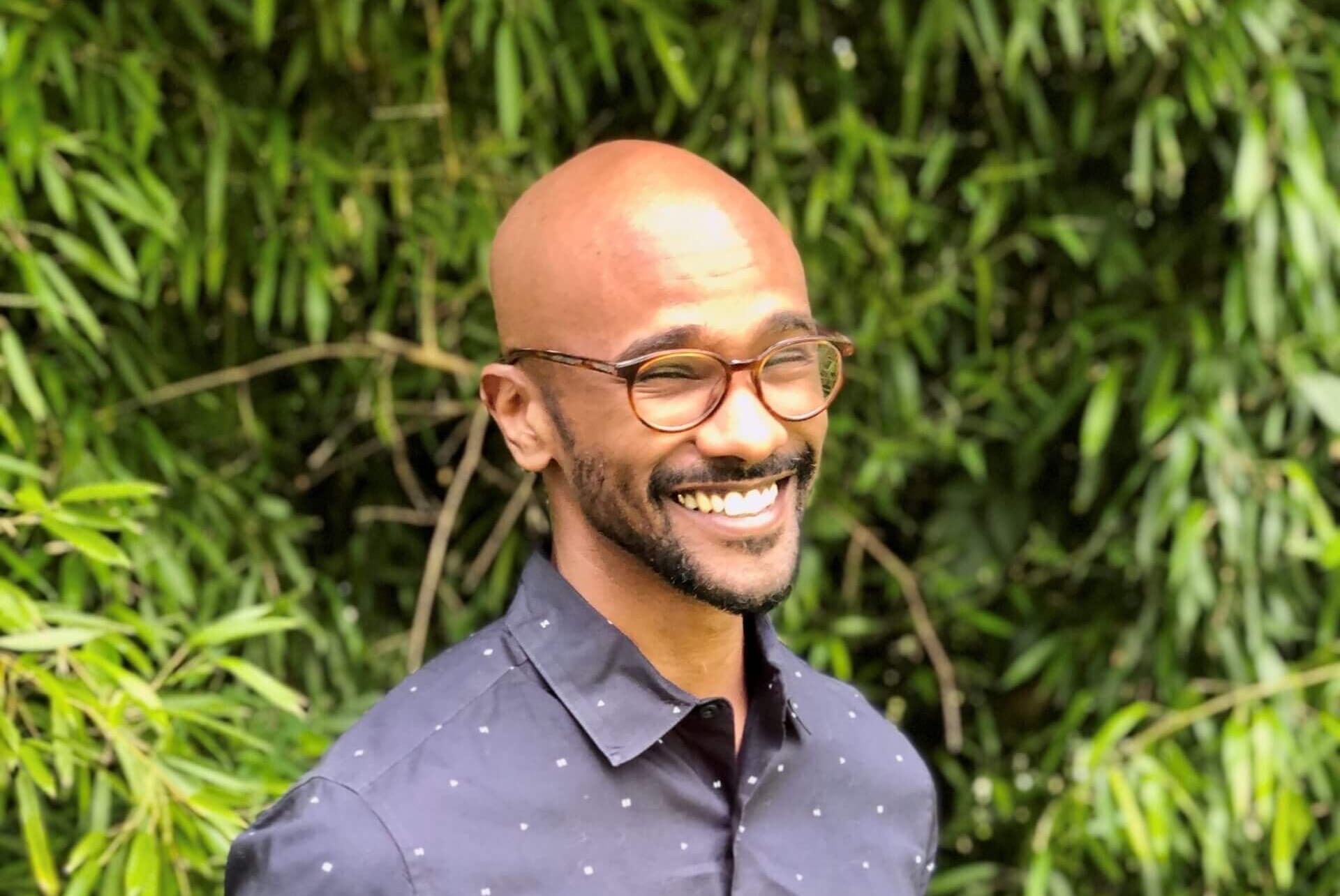 A smiling man, named Sama Ahmed, with glasses and a dark, patterned button-up shirt stands outdoors amid lush green foliage, exuding confidence with his hands in his pockets.