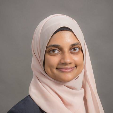 A woman named, Raima Amin, wearing a light pink hijab and a dark top is smiling softly, facing the camera against a plain grey studio background.