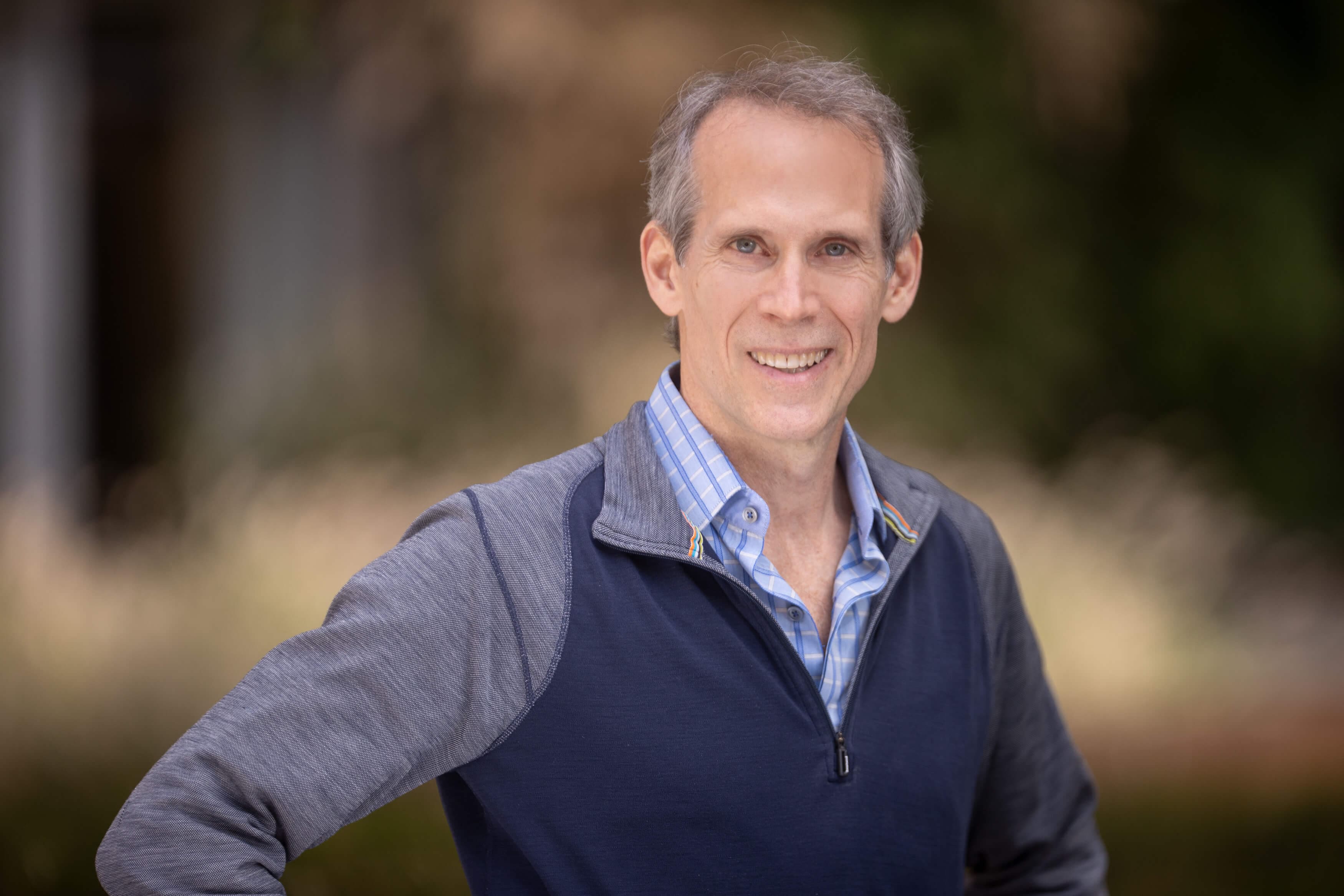 A middle-aged man named Wyeth Bair with short gray hair smiles at the camera. He is wearing a blue zip-up jacket over a blue plaid shirt, standing outdoors in front of a blurred green and brown nature background.