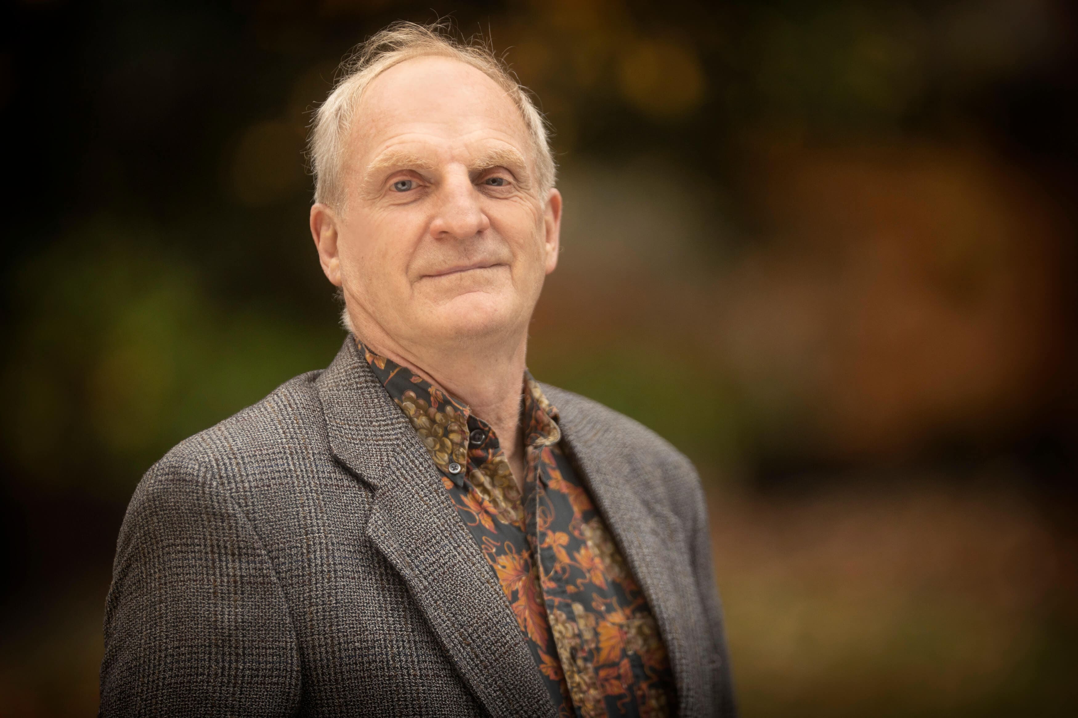 Mark Bothwell, an older man with short gray hair, stands outdoors in a patterned shirt and gray blazer. He looks confidently at the camera with a neutral expression, set against a blurred, natural background.