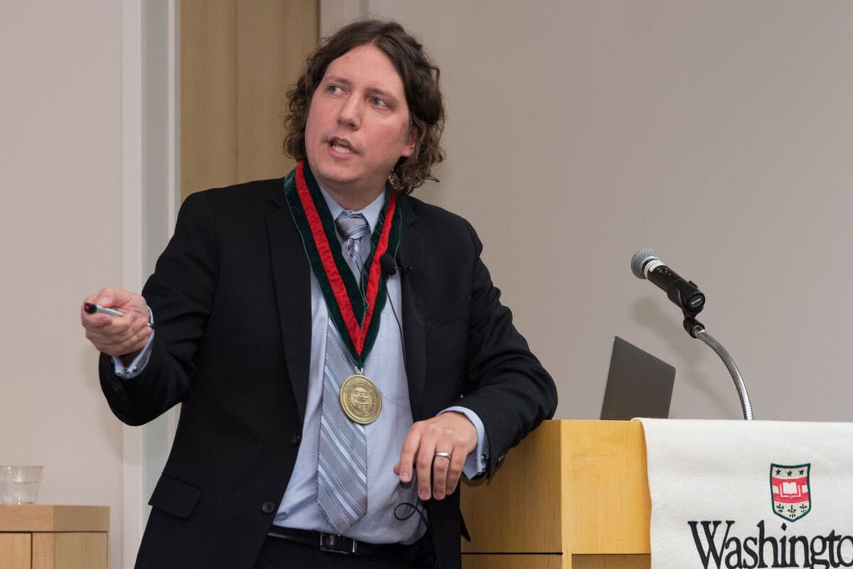 Michael Bruchas, a man with shoulder-length curly hair wearing a suit, medal, and tie, gestures while speaking at a podium with a microphone and a banner with the word 