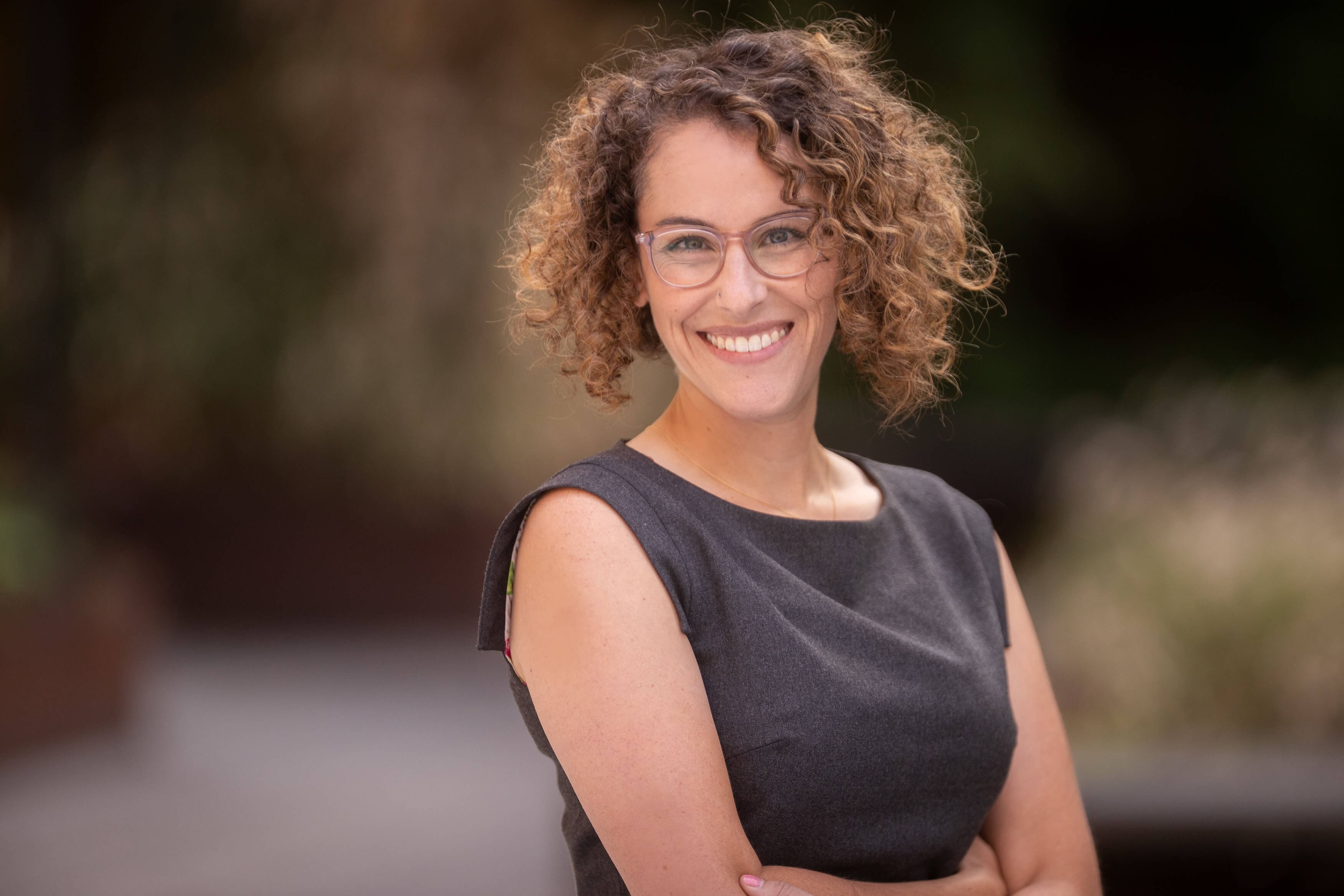 Astra Bryant, a woman with curly hair and glasses, smiles while standing outdoors with her arms crossed. She wears a sleeveless dark gray dress, and the blurred background is filled with greenery.