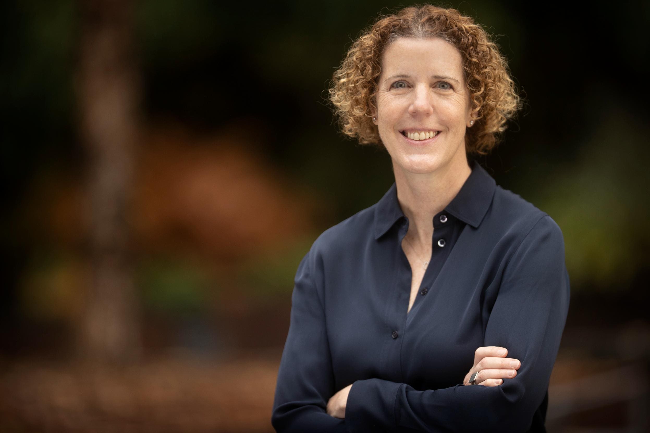 Elizabeth Buffalo, with short curly hair and a dark button-up shirt, stands outdoors with arms crossed and smiles at the camera. The blurred background features earthy tones.