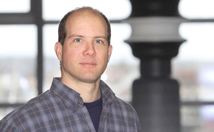 Clemens Cabernard, a man with short hair wearing a plaid shirt, stands indoors in front of a blurred window and column, looking directly at the camera with a neutral expression.