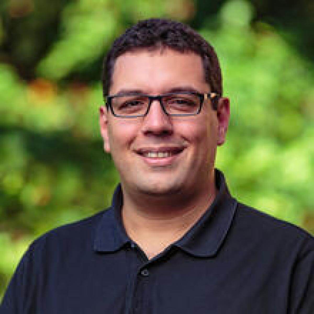 Zaid Harchaoui, a man with short dark hair and glasses, smiles at the camera. He is wearing a dark polo shirt and stands in front of a blurred green outdoor background.