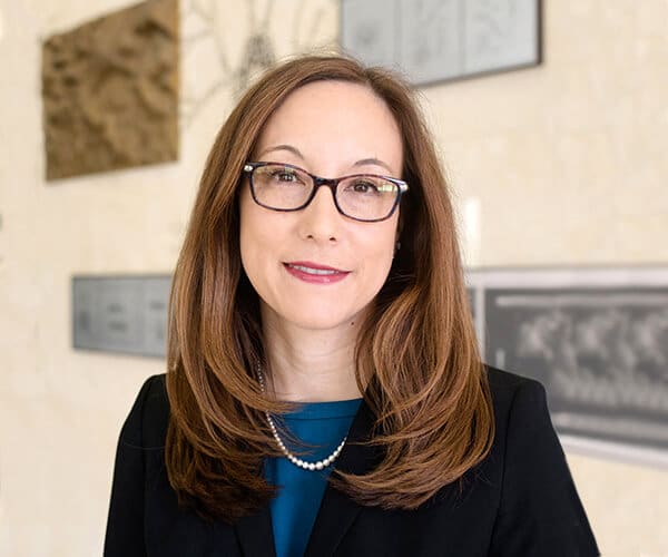 Shelly Sakiyama-Elbert, a woman with long brown hair and glasses, wearing a black blazer, blue top, and pearl necklace, stands indoors in front of a light-colored wall adorned with framed artwork.