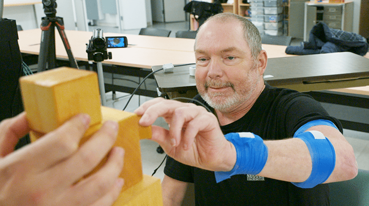 A man with blue medical wraps on his arm, participating in a brain-device trial, reaches out to touch stacked wooden blocks on a table, showcasing fine-motor hand control amid cameras and equipment in the background.