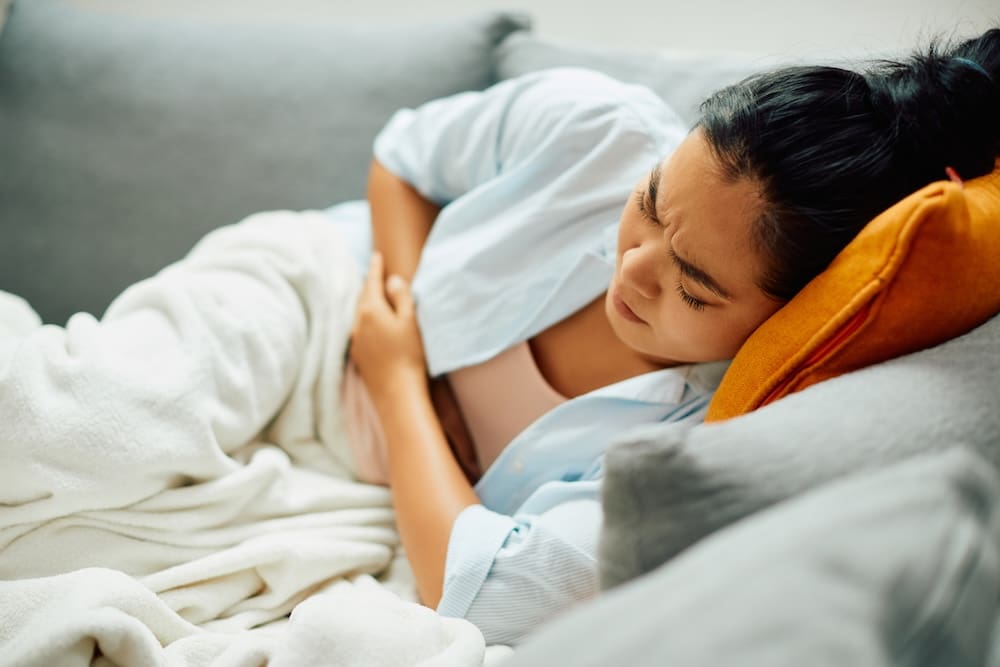 A woman lies on a couch with her eyes closed, holding her stomach in discomfort. Covered with a white blanket and resting on an orange pillow, she appears to be suffering from lasting pain, possibly due to endometriosis.